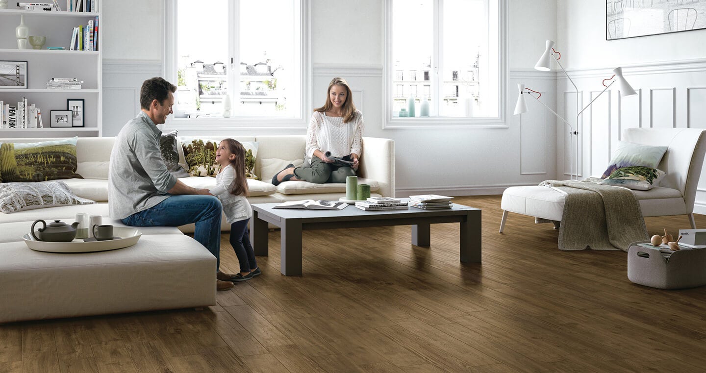 A family including a husband, wife and child are seated in a living room with walnut-colored wood flooring.