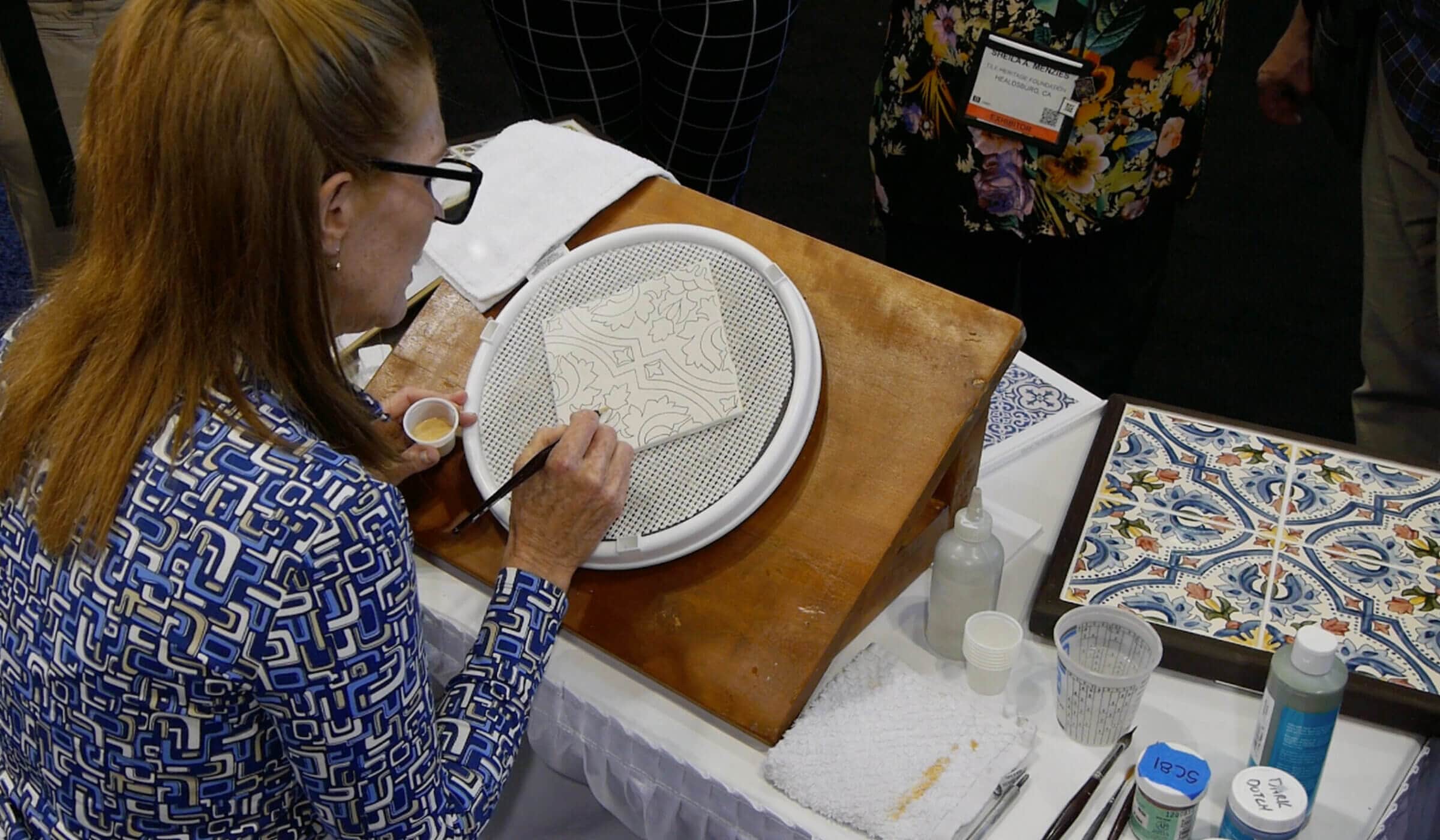 A woman seated at a table, carefully handpainting a tile with an intricate design. 