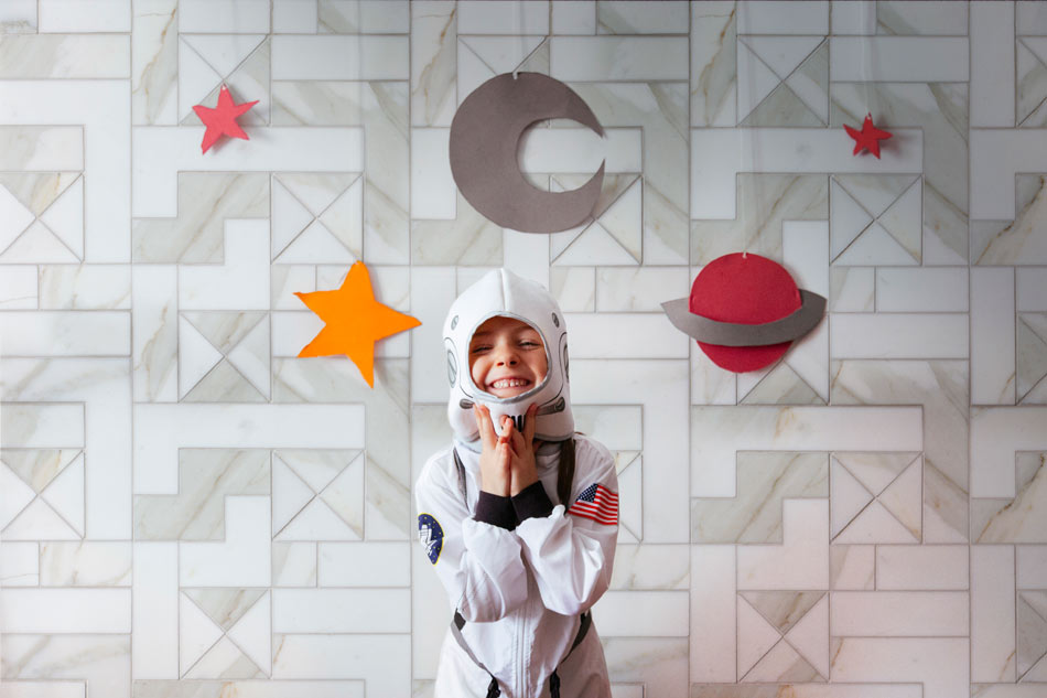 Smiling child in astronaut costume standing in front of marble-look wall tile with custom geometric layout and space-themed decorations