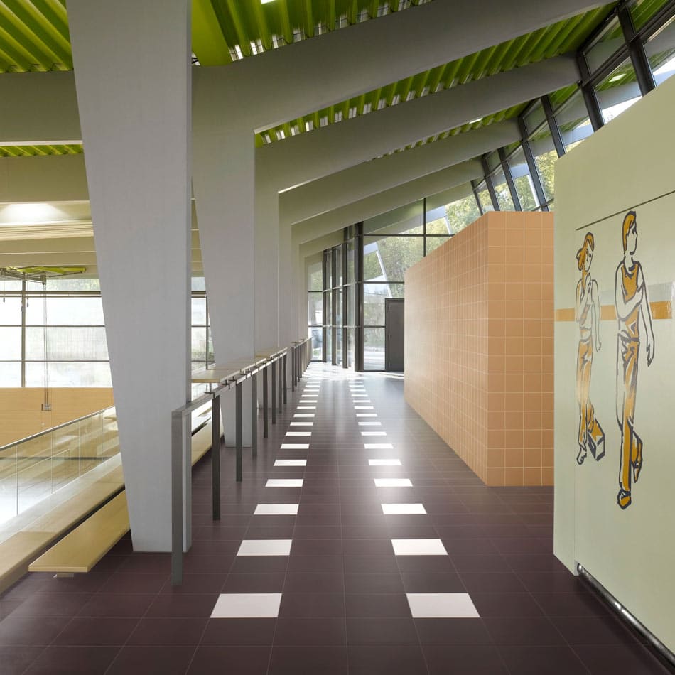 School gym hallway with matte-white and plum floor tiles in a patterned design, apricot tile divider walls, and a mural