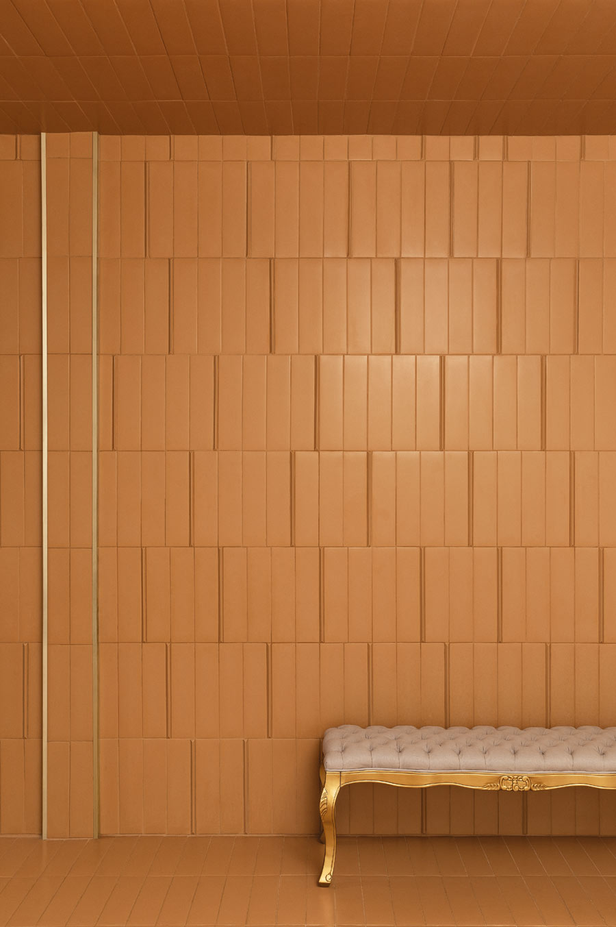 Amber ceramic tile wall and ceiling in a coursing layout with slim vertical pencil tiles and golden trim above a tufted bench