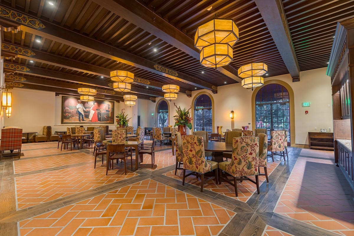 Dining hall with terracotta-look floor tiles, wood-look tile framing a herringbone layout, and a beamed ceiling with double-decker chandeliers